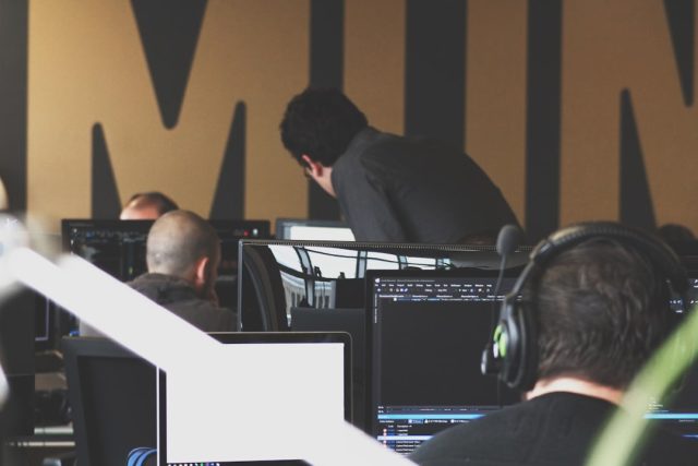 Aura Data Breach Originated From Voice Phishing Call, 900,000 Affected Hacker wearing headset speaks into phone while typing on laptop, digital lock icons float in dark background.